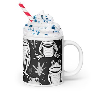 May include: White ceramic mug featuring a black and white frog design. The mug is filled with whipped cream and blue sprinkles. A red and white striped straw protrudes from the whipped cream.