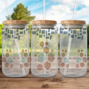 Pode incluir: Três copos de vidro transparentes com tampas de bambu e canudos. Cada copo tem um padrão geométrico de hexágonos marrons, azuis e verdes.