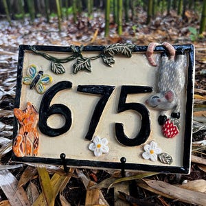 May include: A rectangular ceramic house number plaque featuring the black numbers 675 on a cream background. The plaque is decorated with a sculpted orange cat, a colorful butterfly, green vines, white flowers, and a gray mouse holding a red strawberry.