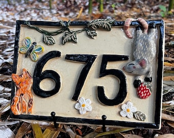 May include: A rectangular ceramic house number plaque displaying the black digits 675 on a cream background. The plaque features a sculpted orange cat, a colorful butterfly, green vines, white flowers, and a gray mouse holding a red strawberry.