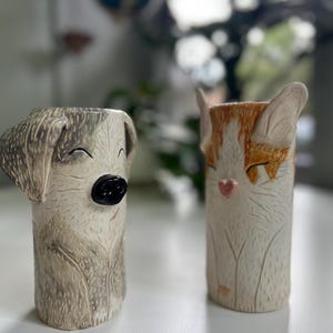 May include: Two cylindrical ceramic vases shaped like animals. The left vase is a grey and white dog with a black nose and closed eyes. The right vase is an orange and white cat with a pink heart-shaped nose and pointed ears, both featuring etched fur textures.