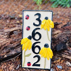 May include: A rectangular ceramic house number plaque featuring the black digits 3662 vertically aligned. The off-white surface is decorated with three yellow coneflower blossoms on green stems and three small red and black ladybug accents.