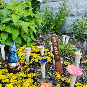 May include: A ceramic gnome figurine with a blue hat and a brown beard, surrounded by small ceramic mushroom garden stakes with colorful tops. A brown sign reads "Bees Please".