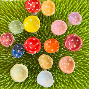 May include: A collection of colorful ceramic mushroom-shaped objects with white polka dots. The mushrooms are in various colors, including red, yellow, orange, pink, and blue. They are arranged on a green, textured surface.