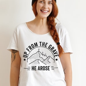 May include: White t-shirt with a black graphic design. The design features a mountain range with three crosses and the text "UP FROM THE GRAVE HE AROSE" in a circular and linear layout. The shirt has a scoop neck.