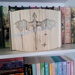 Könnte beinhalten: Eine Buchskulptur mit einem Drachendesign, das in die Seiten geschnitten wurde. Der Drache hat ausgebreitete Flügel und einen langen Schwanz. Der Text "Appleyard" ist sichtbar. Das Buch steht auf einem weißen Regal mit anderen Büchern.