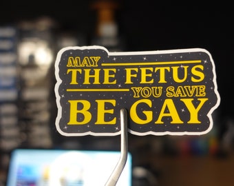 Puede incluir: Una pegatina con el texto "MAY THE FETUS YOU SAVE BE GAY" en amarillo, con fondo negro y estrellas blancas. La pegatina está sujeta por unas pinzas.