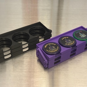 May include: Three round containers with lids are stacked in a purple plastic holder. The lids have labels with the names "Cold Mix", "Black & Cherry", and "California".