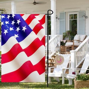 May include: A red, white, and blue American flag garden flag with a white pole. The flag is waving in the wind and is hanging from a white porch railing.