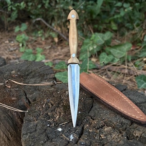 May include: A dagger with a light wood handle and a silver blade is displayed on a tree stump. The dagger is accompanied by a brown leather sheath with a decorative pattern. The background features green foliage.
