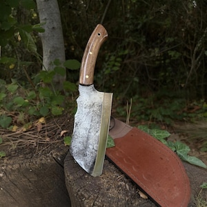 May include: A rustic cleaver knife with a wooden handle and brass rivets. The blade has a weathered, textured appearance. A brown leather sheath is visible next to the knife. The knife is resting on a tree stump.