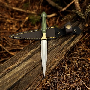 May include: A green and gold dagger with a leather sheath. The dagger is resting on a wooden log with brown and gold dry leaves surrounding it.
