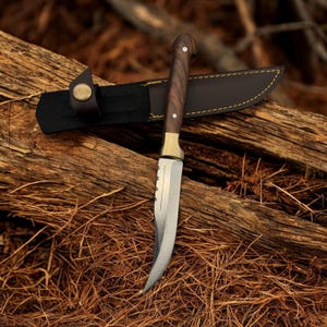 Può includere: Un coltello da caccia ricurvo con manico in legno marrone e fodero in pelle. Il coltello è appoggiato su un mucchio di ramoscelli marroni.