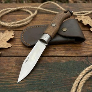 May include: A folding pocket knife with a wooden handle and a stainless steel blade. The knife is open, revealing the sharp edge. It rests on a wooden surface next to a brown leather sheath. A small flower emblem is on the handle.