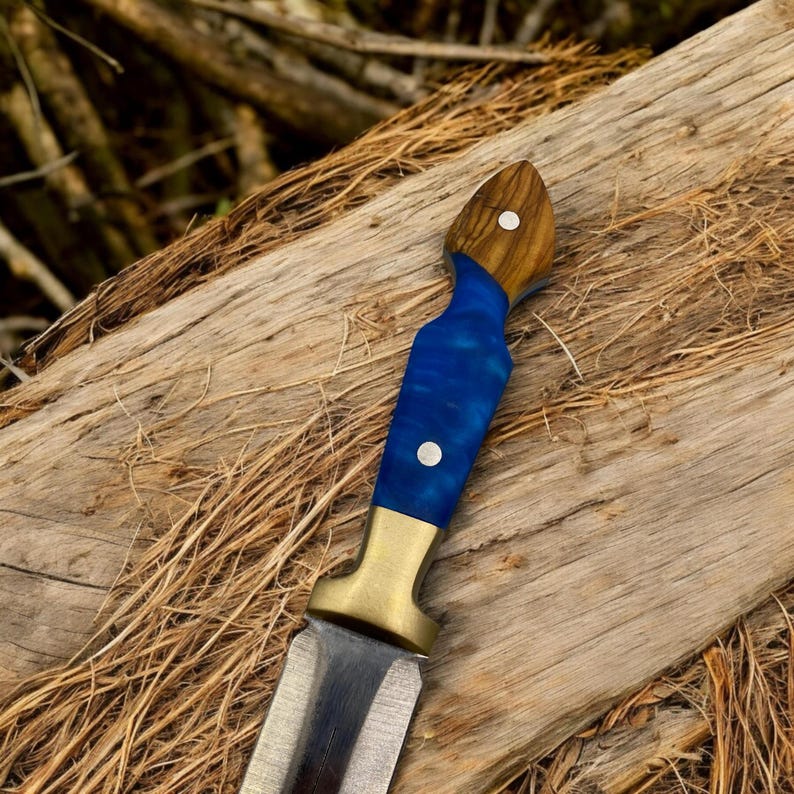 May include: A blue and brown handled knife with a brass guard and a silver blade. The knife is resting on a piece of wood.