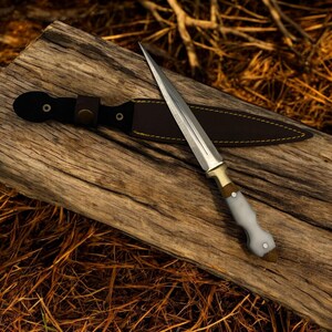 May include: A white-handled dagger with a gold-colored hilt and a brown leather sheath. The dagger is lying on a piece of weathered wood.