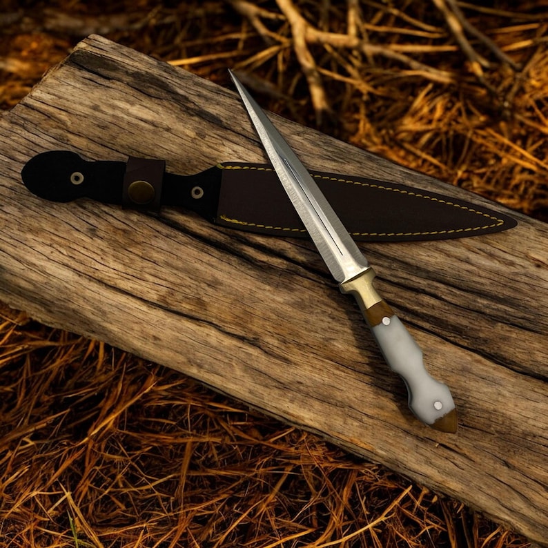 May include: A white-handled dagger with a gold-colored hilt and a brown leather sheath. The dagger is lying on a piece of weathered wood.