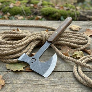 Könnte beinhalten: Eine Stahlaxt mit Holzgriff, die auf einer Holzoberfläche mit einem Seil liegt. Der Axtkopf hat ein Mond- und Stern-Design. Der Griff ist dunkelbraun mit kleinen Nieten.