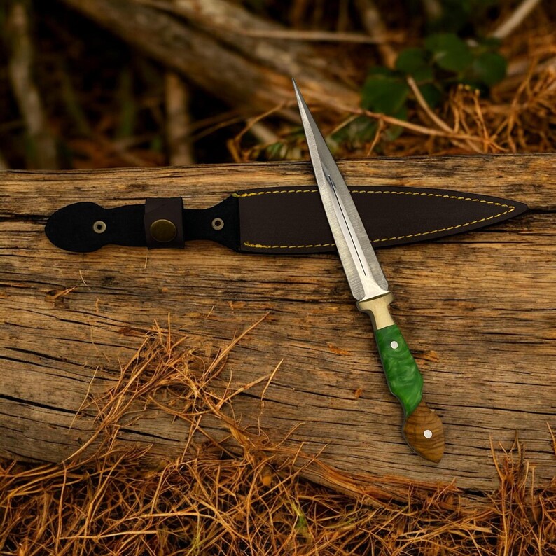 May include: A silver dagger with a green and brown handle and a leather sheath. The dagger is lying on a wooden surface.