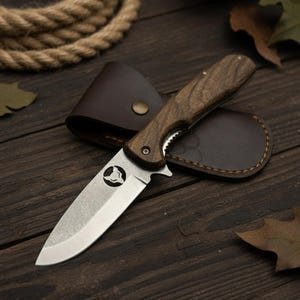 May include: A folding pocket knife with a wooden handle and a stainless steel blade. The knife is resting on a brown leather sheath. The blade has a unique emblem. The knife is approximately 20 cm long when open.