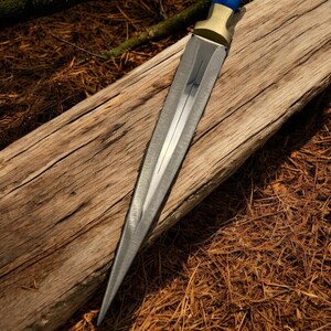 May include: A long, silver dagger with a blue and gold handle. The dagger is lying on a piece of weathered wood.