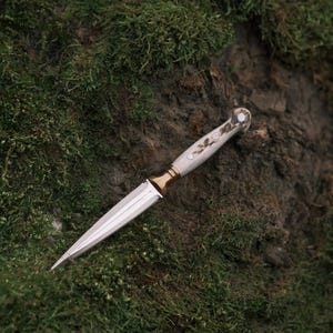 May include: A silver dagger with a white antler handle and gold accents. The dagger is lying on a bed of green moss.