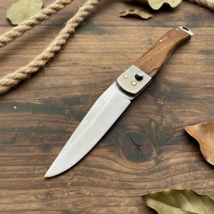 May include: A folding pocket knife with a wooden handle and a silver blade. The knife has a metal bolster with two small rivets. The handle is curved and appears to be made of a dark wood. The knife is on a wooden surface.