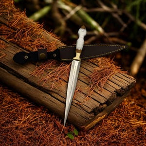 May include: A silver dagger with a white handle and a leather sheath. The dagger is resting on a piece of wood with brown bark.