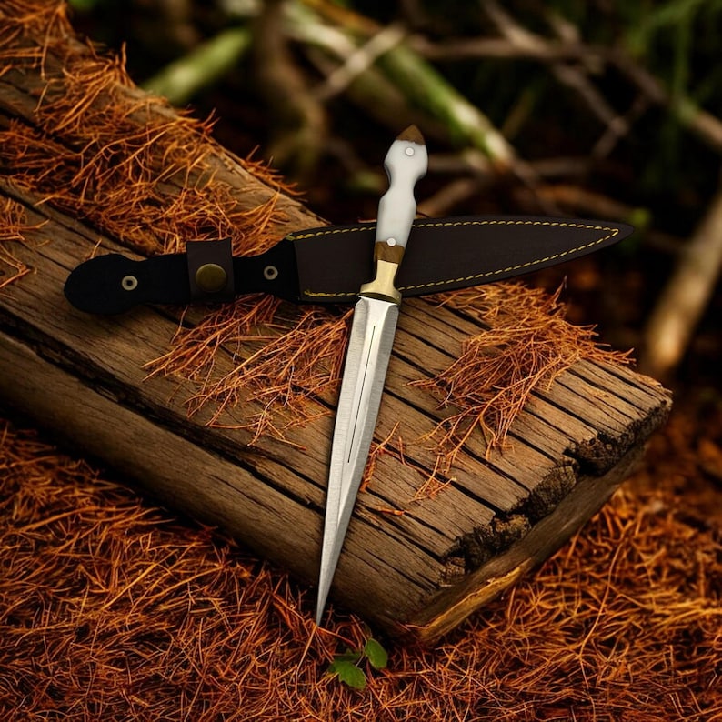 May include: A silver dagger with a white handle and a leather sheath. The dagger is resting on a piece of wood with brown bark.