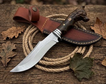 May include: A fixed-blade knife with a silver steel blade and a dark brown handle carved into the shape of a wolf head. The knife includes a brown leather sheath with a snap closure and decorative stitching, resting on a coiled tan rope.