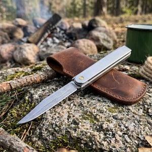 May include: A silver folding knife with a patterned blade rests in a brown leather sheath. The knife is open, revealing the blade's design. The scene is set outdoors, with a campfire and green enamel mug in the background, suggesting a camping or outdoor theme.
