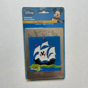 May include: A silver metal die cut for a craft project featuring a pirate ship with Mickey Mouse's head on the sail. The die cut is packaged in a clear plastic sleeve with a Disney logo and the text "Ahoy Matey Ohé les Copains cut découpe 1 cutting die 1 matrice de découpe".