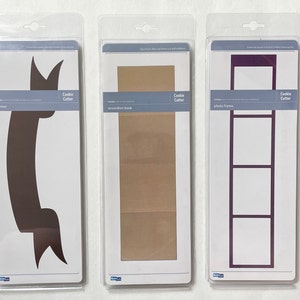 May include: Three cookie cutter templates packaged in clear plastic. The cookie cutters are for a banner, an accordion book, and a photo frame. The cookie cutters are all brown and purple.