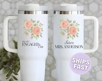 May include: White insulated tumbler with a handle, featuring a floral design of peach-colored roses and green leaves. Text on the tumbler reads "IN MY ENGAGED era" and "Future MRS. ANDERSON". A purple sticker with "SHIPS FAST" is also visible.