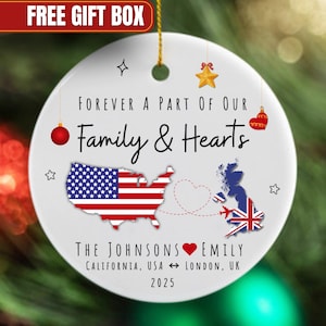 May include: A white ceramic Christmas ornament with a gold string. The ornament features the text "Forever a part of our Family & Hearts" and a map of the USA and UK with flags. The names "The Johnsons Emily" and the year 2025 are also included.