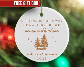 May include: White ceramic Christmas ornament with gold string. The ornament features the text "A FRIEND IS GOD'S WAY OF MAKING SURE WE never walk alone" with two pine trees and the names "Ashley & Jessica 2025".