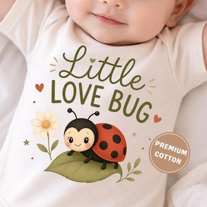 May include: A cream-colored baby bodysuit featuring a graphic of a ladybug on a green leaf next to a daisy. The text reads Little LOVE BUG in dark green letters. A circular badge on the lower right side contains the text PREMIUM COTTON.