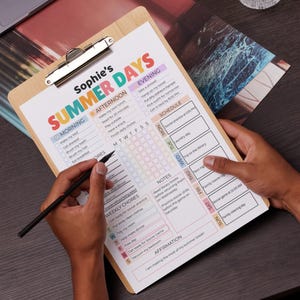 May include: A wooden clipboard with a printed summer schedule titled "Sophie's SUMMER DAYS." The schedule includes sections for morning, afternoon, and evening activities, weekly chores, and a schedule. A hand holds a pen over the schedule.