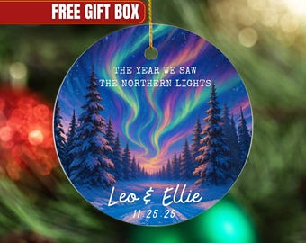 May include: Round Christmas ornament featuring a vibrant depiction of the Northern Lights over a forest. Text reads "The Year We Saw The Northern Lights" with names "Leo & Ellie" and date "11.25.25". Includes a gold string for hanging.