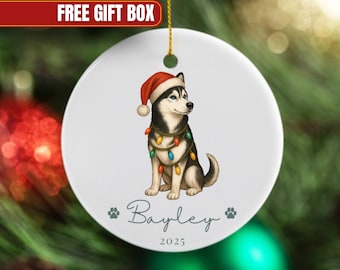May include: A white ceramic Christmas ornament featuring a Siberian Husky wearing a Santa hat and string lights. The ornament has the name "Bayley" and the year "2025" printed on it. The words "FREE GIFT BOX" are in a red banner.