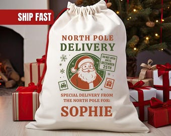 May include: A large, off-white Christmas gift bag with a drawstring closure. The bag features the text "NORTH POLE DELIVERY" and a Santa Claus graphic. The name "SOPHIE" is printed below. Presents and a Christmas tree are in the background.