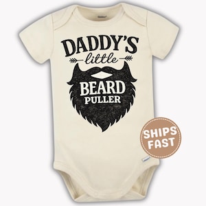 May include: Cream-colored baby bodysuit with the text "DADDY'S little BEARD PULLER" in black. The design includes a stylized beard graphic. The bodysuit has short sleeves and a round neckline.
