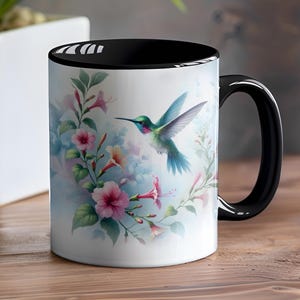 Puede incluir: Taza de cerámica blanca con interior y asa negros. La taza presenta un diseño de acuarela de un colibrí en vuelo, rodeado de flores rosas y melocotón y hojas verdes. El fondo es azul claro.