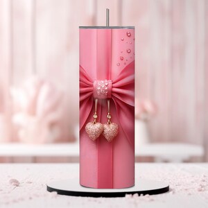 Pu&ograve; includere: Un bicchiere in acciaio inossidabile rosa con un fiocco rosa e due ciondoli a forma di cuore dorati. Il bicchiere ha una finitura lucida e metallica.