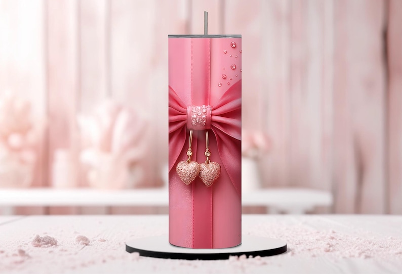 Pu&ograve; includere: Un bicchiere in acciaio inossidabile rosa con un fiocco rosa e due ciondoli a forma di cuore dorati. Il bicchiere ha una finitura lucida e metallica.