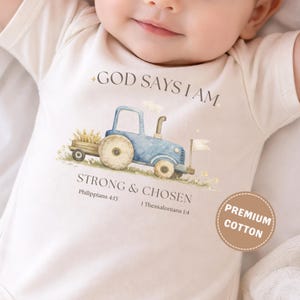 May include: Cream-colored baby onesie with a blue tractor illustration and the text "GOD SAYS I AM STRONG & CHOSEN." The onesie also has a "PREMIUM COTTON" label. The design features wheat and a white flag.