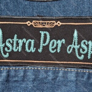 May include: A rectangular black embroidered patch featuring the Latin text Ad Astra Per Aspera in light blue cursive lettering. The patch is bordered by a thin tan line with decorative corner accents and is attached to a blue denim fabric surface.