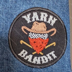 May include: A circular embroidered patch with a black background and a stitched border. The design features a brown cowboy hat above a red bandana and two crossed knitting needles. The white text reads YARN at the top and BANDIT at the bottom.
