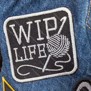 May include: A square embroidered patch with rounded corners attached to blue denim fabric. The patch features a black background with white stitched text that reads WIP LIFE and a white yarn ball graphic with a knitting needle.