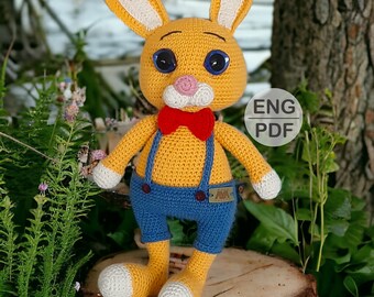May include: A crocheted yellow bunny rabbit wearing blue overalls and a red bow tie. The bunny has large, black eyes and a pink nose. The bunny is sitting on a wooden surface with green foliage in the background. ENG PDF is written on a white tag.